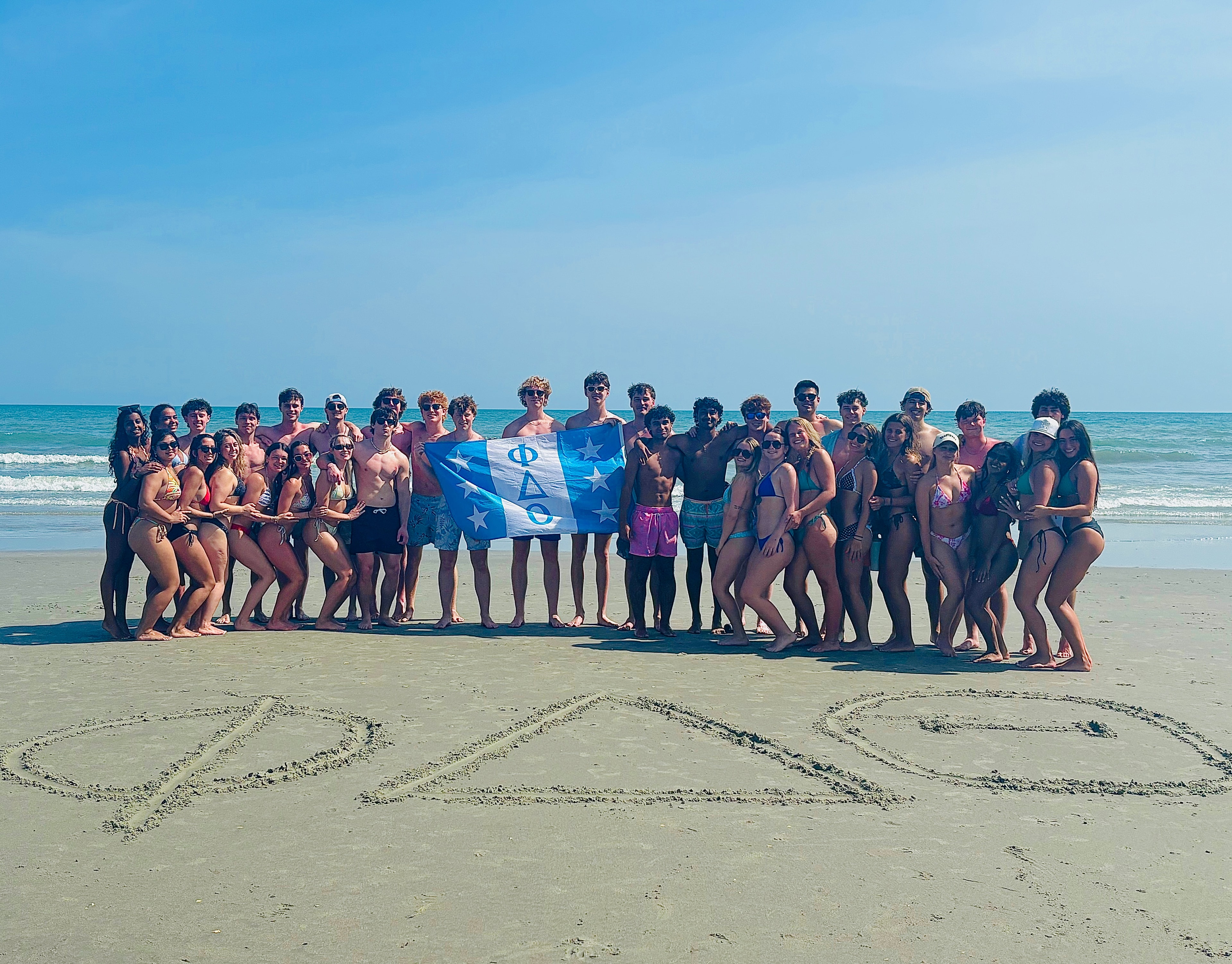 Brothers of NC Epsilon on the beach holding the chapter flag, with the Greek letters drawn in the sand in front of them