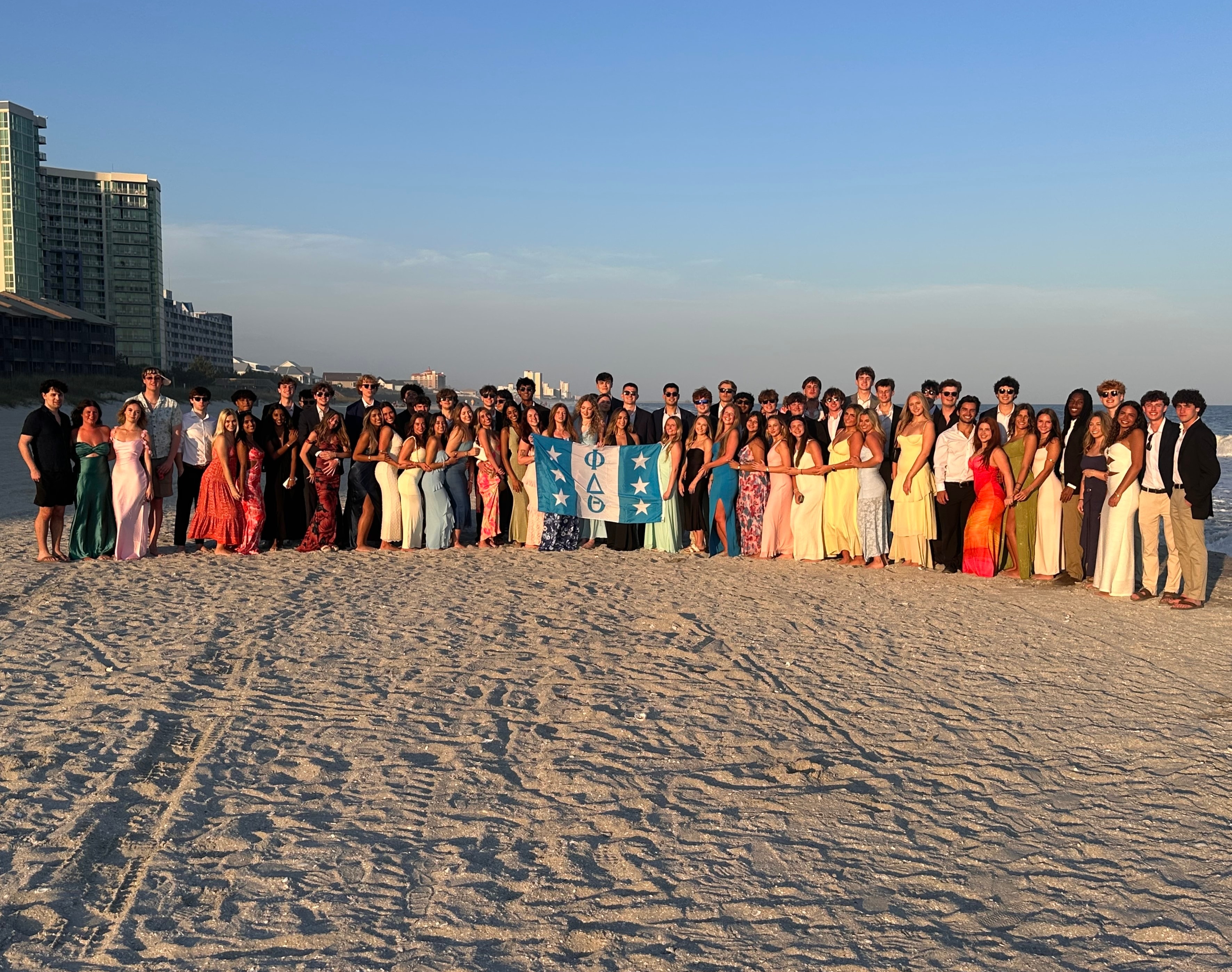 NC Epsilon brothers and their dates in formal wear on the beach at golden hour, gathered around the chapter flag