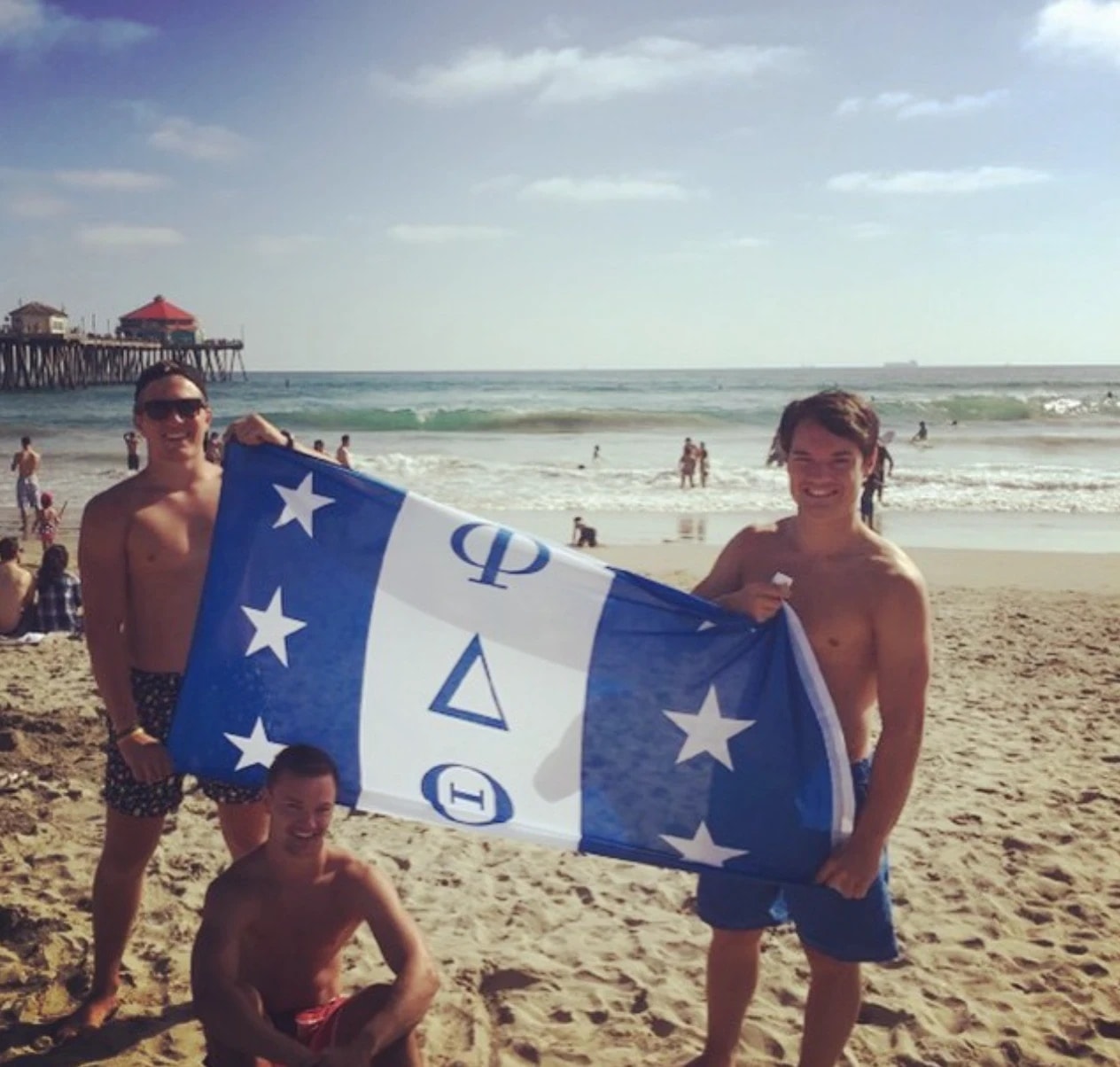 Three brothers on the beach holding the Phi Delta Theta flag, with a pier and the ocean behind them